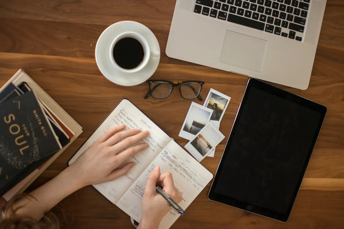 Schreibtisch mit Notizbuch, Kaffee, Laptop und Tablet von oben.